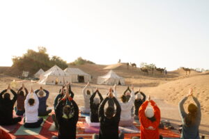 Groupe de participants pratiquant le yoga dans un campement du désert entouré de dunes et de dromadaires.